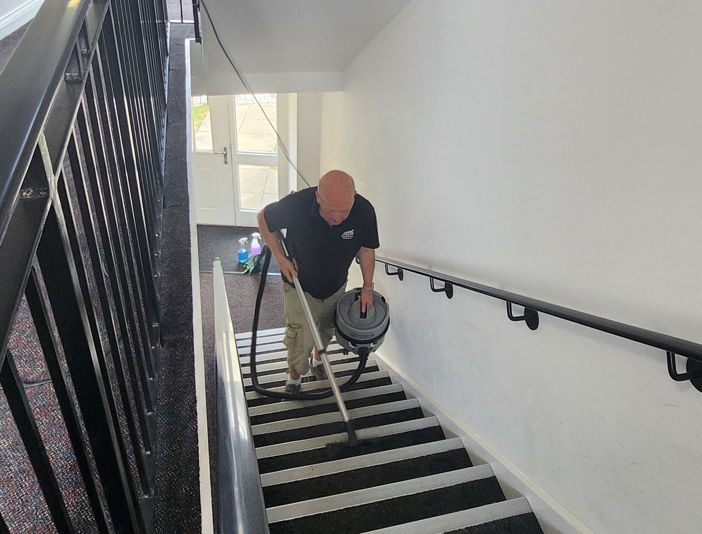 A male hoovering the stairs in a communal area in a block of flats to stop complaints about lack of cleanliness.