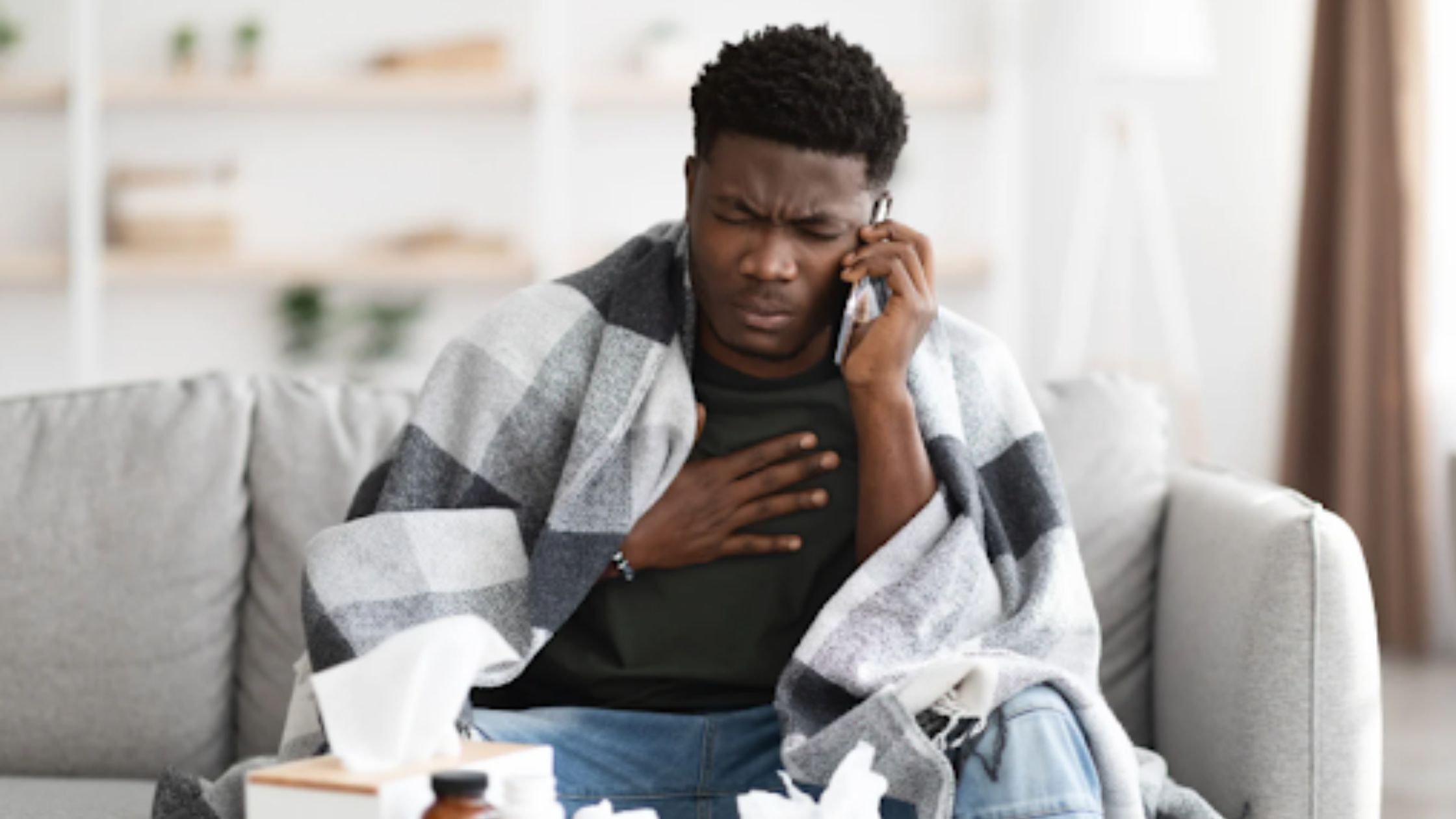 Unwell office worker wrapped in a blanket, holding his chest while talking on the phone, highlighting the impact of poor office cleaning on employee health.